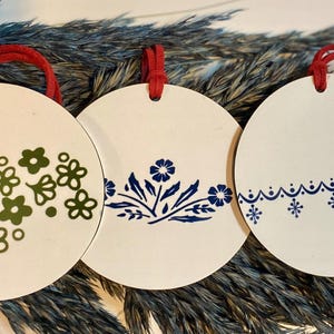 May include: Three white, round ornaments with red hanging loops. Each ornament features a different floral design in green or blue. The ornaments are displayed against a backdrop of dried foliage.