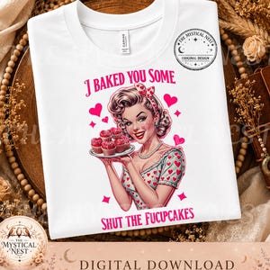 May include: A white t-shirt featuring a graphic of a person with styled hair holding a plate of cupcakes. The text reads "I BAKED YOU SOME" and "SHUT THE FUCUPCAKES" in pink letters, surrounded by pink hearts and a circular logo.