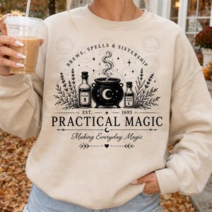 Puede incluir: Una sudadera beige de cuello redondo con un estampado gráfico negro de un caldero, dos frascos de pociones y ramas de lavanda. El texto dice: BREWS, SPELLS & SISTERSHIP, EST. 1693, PRACTICAL MAGIC y Making Everyday Magic con pequeñas estrellas.