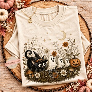 A beige crewneck t-shirt with a vintage-style Halloween illustration featuring a black cat in a witch hat, three ghosts, a jack-o-lantern, flowers, and stars.
