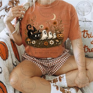 Rust-colored t-shirt featuring whimsical Halloween cat with witch hat, three ghosts, pumpkin, mushrooms, moon, and botanical autumn artwork for apparel mockup.