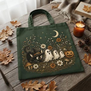 A dark green tote bag featuring the vintage black cat and ghost Halloween artwork resting on a wooden table with autumn decor.