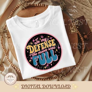 Op de afbeelding: Een wit t-shirt met een cirkelvormige afbeelding op een zwarte achtergrond, roze rand en geel-blauwe tekst met de tekst In my DEFENSE the moon was FULL, omringd door kleine gele en roze sterillustraties.