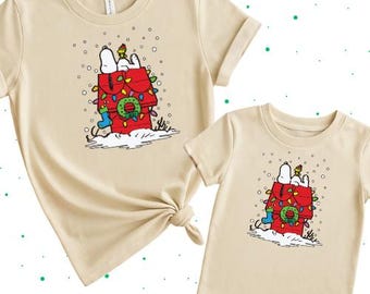 May include: Two beige t-shirts, one adult and one child size, featuring a Snoopy graphic. The graphic shows Snoopy on top of his red doghouse decorated with Christmas lights and a wreath. Snowflakes are falling around the doghouse.