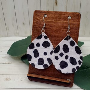May include: A pair of teardrop-shaped earrings with a white background and black spots, resembling a cowhide pattern. The earrings are displayed on a wooden stand with silver-colored hooks.