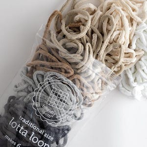 May include: A clear plastic bag filled with assorted cotton loops for weaving, featuring shades of white, beige, tan, grey, and dark blue. The packaging is labelled with the text friendly loom, traditional size, lotta loops, and makes 6 potholders.