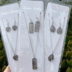 May include: Three sets of fashion jewellery, each including a necklace and matching earrings. The necklaces have silver chains with pendants in heart, rectangular, and teardrop shapes. The earrings are also silver with matching pendant shapes. The jewellery is packaged on white cards.