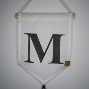 Könnte beinhalten: Weißer Canvas-Banner mit einem schwarzen Buchstaben M darauf gedruckt. Der Banner hängt an einer Holzleiste mit einem weißen Seil. Ein schwarzer Quaste hängt am unteren Ende des Banners.