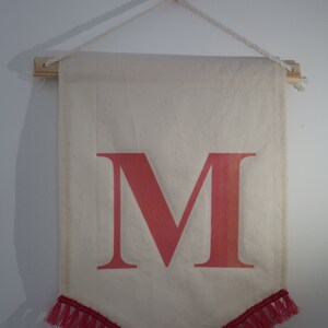 Könnte beinhalten: Ein weißes Canvas-Banner mit einem großen, rosafarbenen Buchstaben "M" darauf gedruckt. Das Banner hat einen rosafarbenen Fransenrand am unteren Rand und hängt an einem weißen Seil mit einem rosafarbenen Knopf.