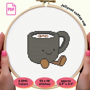 Peut inclure: Un motif de point de croix d'une tasse à café grise souriante avec des grains de café bruns, maintenu dans un cercle à broder en bois. Le texte "Jellycat coffee cup" est au-dessus du motif. Le motif mesure environ 9,9 cm x 8,6 cm.