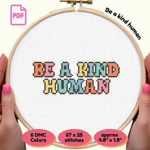 May include: A cross-stitch design in a wooden hoop, featuring the phrase "BE A KIND HUMAN" in colorful, pixelated letters. The design is held by hands with pink nail polish. The text "Be a kind human" is written above the design.