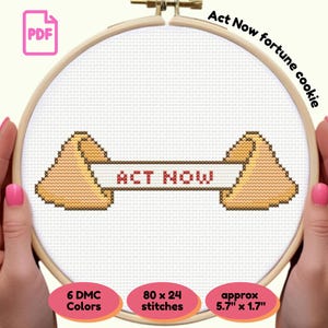 Op de afbeelding: Kruissteekpatroon van een gelukskoekje met de tekst "ACT NOW" op een witte achtergrond, gehouden in een houten borduurring. Het ontwerp bevat 6 DMC-kleuren, 80 x 24 steken en ongeveer 14.5 cm x 4.3 cm.