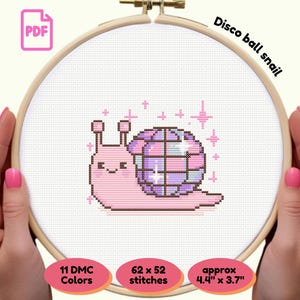 May include: Cross-stitch pattern of a pink snail with a disco ball shell, held in a wooden hoop. The text "Disco ball snail" is above the design. The pattern is approximately 4.4" x 3.7" and uses 11 DMC colors.
