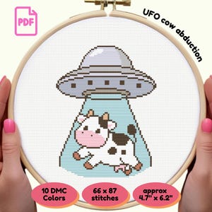 May include: A cross-stitch pattern in a wooden hoop shows a cartoon UFO abducting a cow. The UFO is grey, and the cow is white with black spots. The text "UFO cow abduction" is above the design. The pattern is approximately 12 cm x 16 cm.
