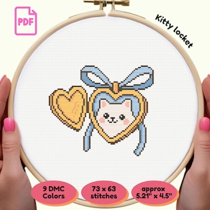 Op de afbeelding: Kruissteekpatroon met een kat in een hartvormig medaillon met een blauwe strik, gehouden in een houten borduurring. Het ontwerp bevat een gouden hart en de tekst "Kitty locket". Het patroon is ongeveer 13,23 cm x 11,43 cm groot.