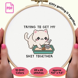 Może przedstawiać: Wzór do haftu krzyżykowego przedstawiający kreskówkowego kota w kuwecie, z napisem "TRYING TO GET MY SHIT TOGETHER". Wzór znajduje się w drewnianym tamborku, z napisem "Kitty getting it together" powyżej. Wzór ma wymiary około 11,7 cm x 11,7 cm.