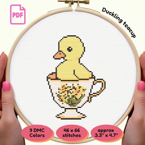 May include: A cross-stitch pattern of a yellow duckling in a teacup, held in a wooden embroidery hoop. The teacup is decorated with yellow flowers. The text "Duckling teacup" is above the design. The pattern is approximately 3.3" x 4.7".