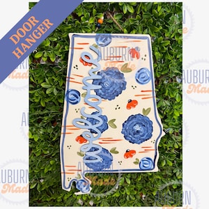 May include: A wooden door hanger shaped like the state of Alabama, featuring a cream background with painted dark blue flowers, small orange accents, and horizontal orange lines. The word Welcome is written in a light blue script font.