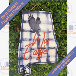 May include: Wooden door hanger in the shape of Alabama, featuring a gray eagle and the words "War Eagle" in orange. The background has a blue and white plaid pattern. "Auburn University Licensed" is printed on a blue banner.