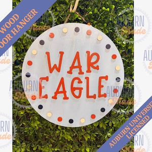 May include: A round, light gray wooden door hanger with the words "WAR EAGLE" in orange. The sign is decorated with small, colorful dots around the edge and includes the text "WOOD DOOR HANGER" and "AUBURN UNIVERSITY LICENSED".