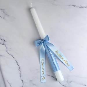 Peut inclure: Une bougie blanche ornée d'un ruban et d'un nœud bleu clair. Le ruban porte l'inscription dorée "Baptême Aline" et "10 Juin 2026" avec un symbole de colombe. Posée sur une surface en marbre.