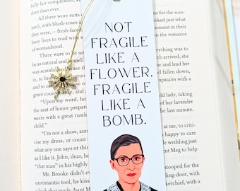 May include: White bookmark with the text "NOT FRAGILE LIKE A FLOWER. FRAGILE LIKE A BOMB." and a portrait of Ruth Bader Ginsburg. The bookmark is hanging over an open book.