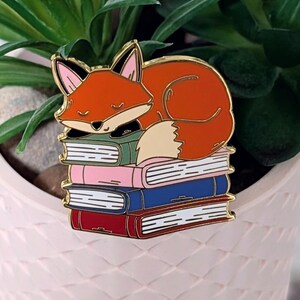 May include: A colorful enamel pin featuring a sleeping fox atop a stack of books. The fox is orange with a white belly and black accents. The books are in shades of green, pink, blue, and red. The pin has a gold outline.
