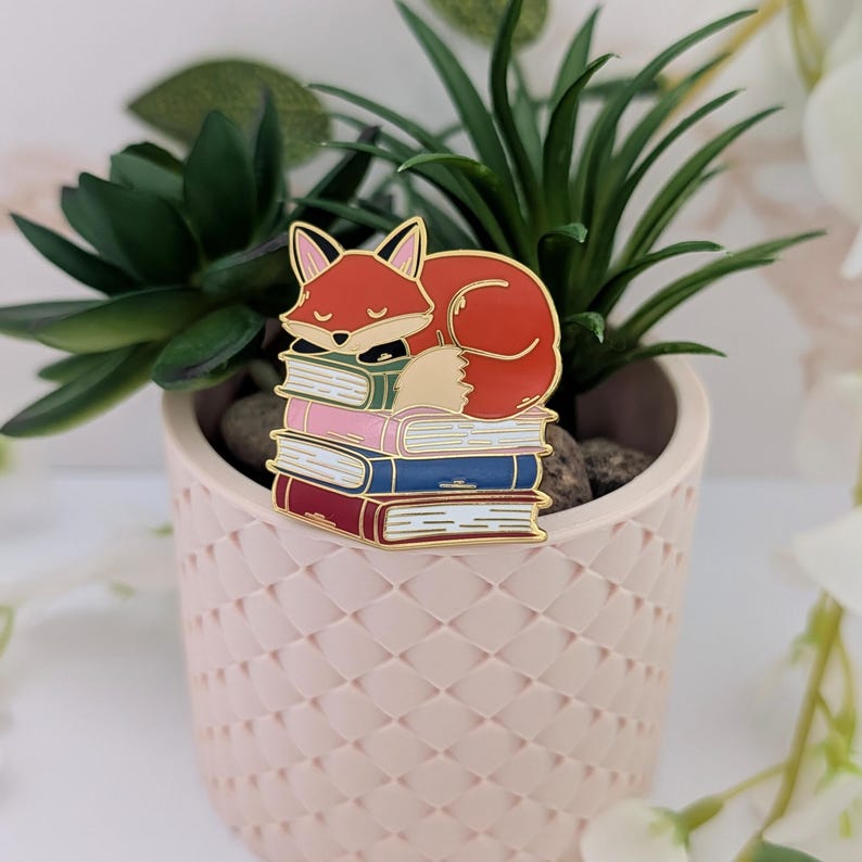 May include: A gold-outlined enamel pin featuring a sleeping fox atop a stack of books. The fox is orange with a white belly and black accents. The books are pink, blue, and burgundy with gold page edges. The pin is set against a blurred background.