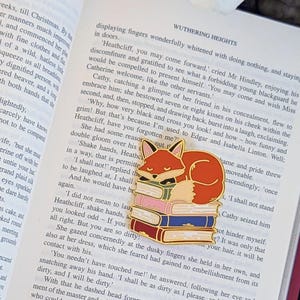 May include: A gold-colored enamel pin featuring a sleeping fox atop a stack of colorful books. The fox is orange and white, with a black face. The books are red, pink, and blue. The pin is set on an open book.