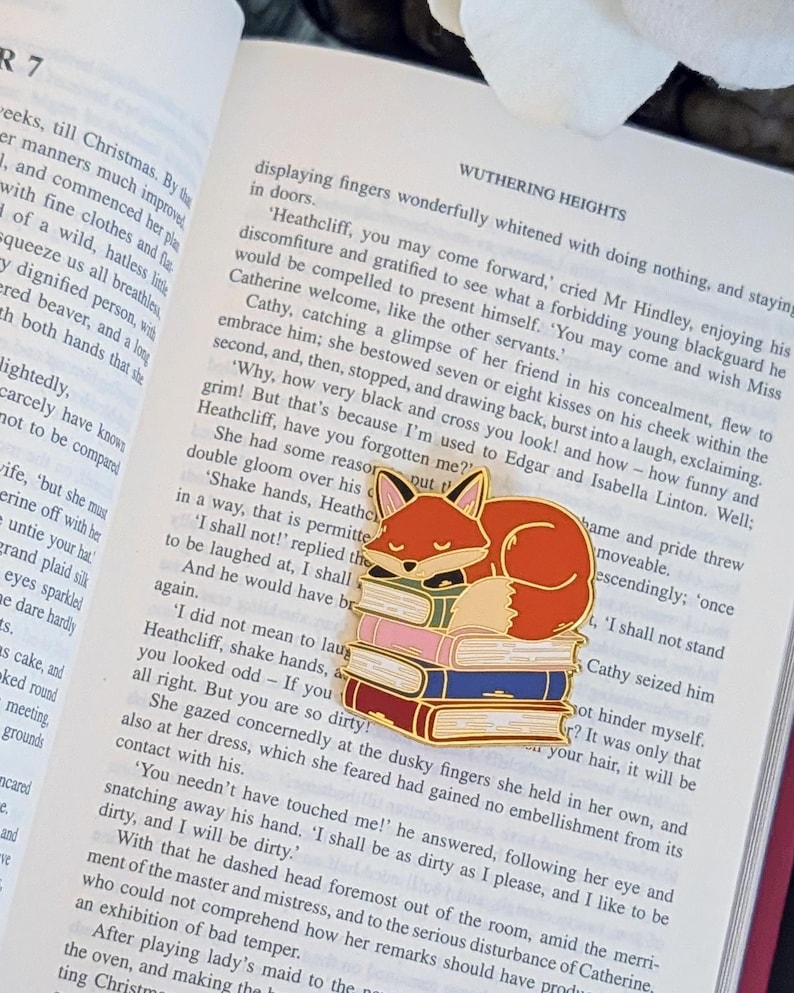 May include: A gold-colored enamel pin featuring a sleeping fox atop a stack of colorful books. The fox is orange and white, with a black face. The books are red, pink, and blue. The pin is set on an open book.