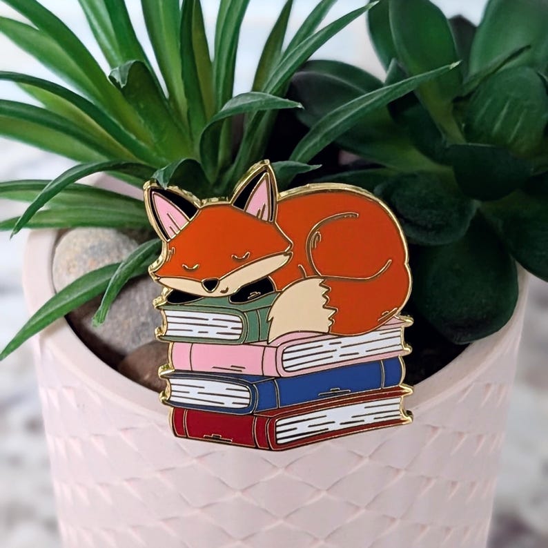 May include: A colorful enamel pin featuring a sleeping fox atop a stack of books. The fox is orange with a white belly and black accents. The books are in shades of green, pink, blue, and red. The pin has a gold outline.