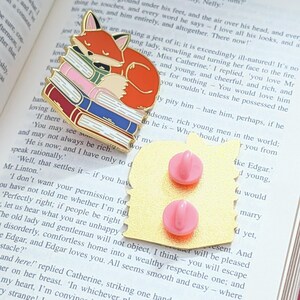 May include: A gold-colored enamel pin featuring a sleeping orange fox atop a stack of colorful books. The pin has two pink pin backs. The fox is curled up, and the books are in shades of red, pink, and blue.