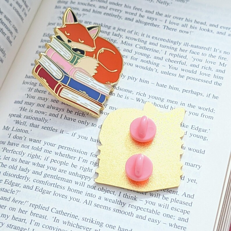 May include: A gold-colored enamel pin featuring a sleeping orange fox atop a stack of colorful books. The pin has two pink pin backs. The fox is curled up, and the books are in shades of red, pink, and blue.