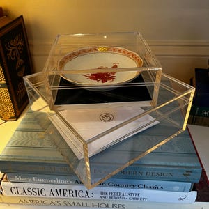 May include: A clear acrylic display box with a decorative plate inside. The plate has a white background with red and gold accents. The box is stacked on top of a stack of books with titles like "Classic America" and "Modern Design".