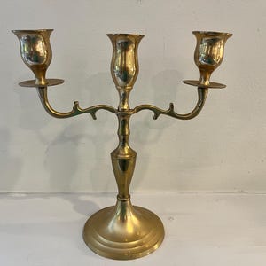 May include: A polished brass candelabra featuring a central stem and two symmetrical arms, each topped with a cup-shaped candle holder. The base is circular and tiered, supporting the vertical structure with a smooth, reflective metallic finish.