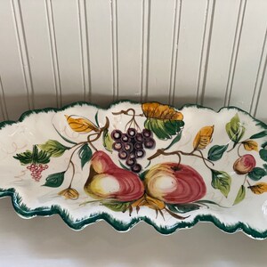 May include: A white ceramic serving dish shaped like a leaf with green trim. The dish features a painted design of fruit, including pears, apples, and grapes, with green leaves and vines.