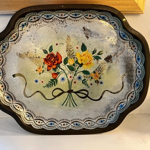 May include: An oval metal serving tray with a dark brown rim and a light green center. The tray features a central floral bouquet of red and yellow roses tied with a black ribbon, surrounded by a decorative border of blue and green patterns.