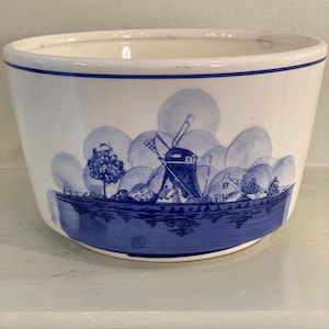 May include: A white ceramic planter with a blue and white windmill scene. The planter has a rounded bottom and a wide rim.