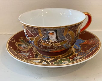 Könnte beinhalten: Ein antikes japanisches Tee-Set mit Tasse und Untertasse mit aufwendigen goldenen und roten Designs. Die Tasse zeigt ein gemaltes Bild einer Frau in traditioneller japanischer Kleidung.