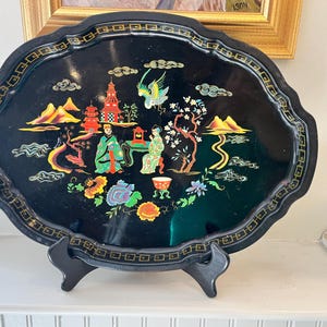 May include: An oval black lacquer tray featuring a colorful hand-painted scene of two figures, a pagoda, mountains, a bird, and floral motifs. The tray has a scalloped edge with a gold geometric border and rests on a black plastic display stand.
