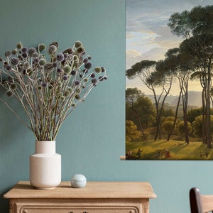 May include: A dried flower arrangement in a white vase sits on a wooden table in front of a painting of a forest scene with tall pine trees. The painting is framed and hanging on a teal wall.