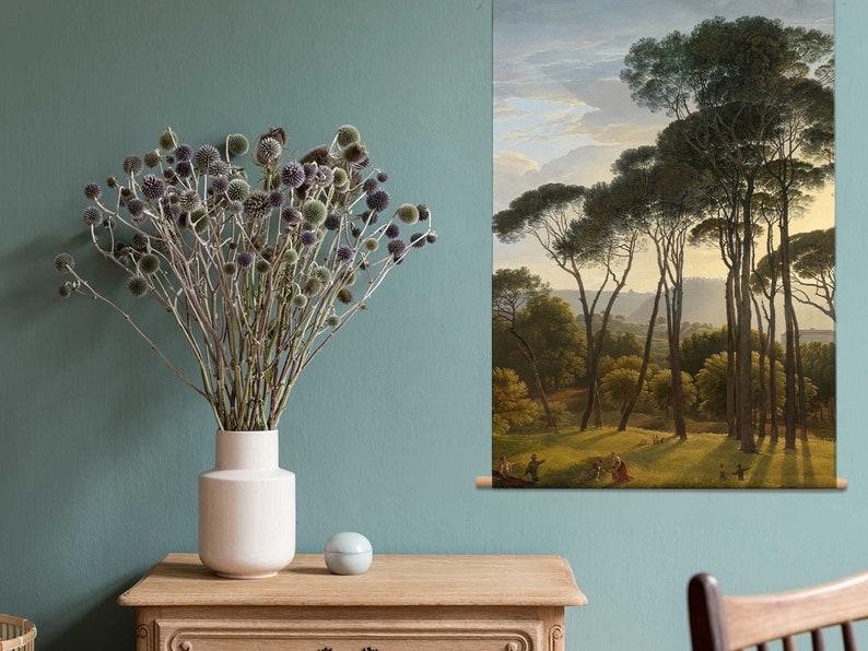 May include: A dried flower arrangement in a white vase sits on a wooden table in front of a painting of a forest scene with tall pine trees. The painting is framed and hanging on a teal wall.