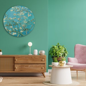 May include: A round wall art piece featuring a painting of a blooming almond tree with white blossoms against a blue sky. The art is hanging on a teal wall in a living room setting with a wooden cabinet, a pink armchair, and a small table with plants.