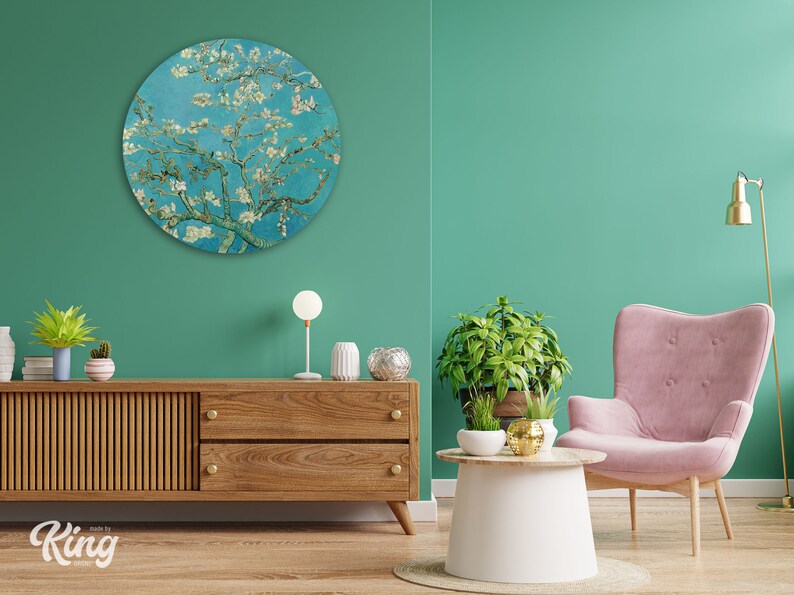 May include: A round wall art piece featuring a painting of a blooming almond tree with white blossoms against a blue sky. The art is hanging on a teal wall in a living room setting with a wooden cabinet, a pink armchair, and a small table with plants.