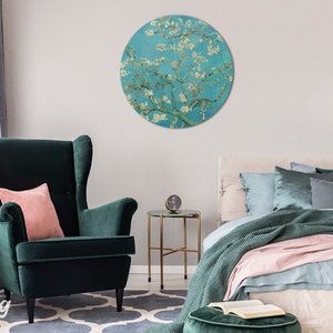 May include: A round wall art print depicting a painting of a blossoming almond tree with white flowers against a blue sky. The print is displayed in a living room setting with a green armchair and a bed with pink and green bedding.