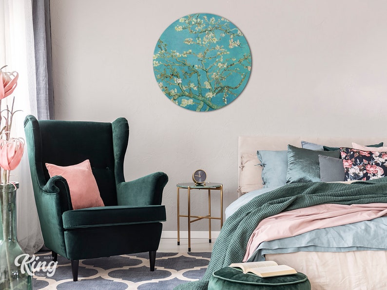 May include: A round wall art print depicting a painting of a blossoming almond tree with white flowers against a blue sky. The print is displayed in a living room setting with a green armchair and a bed with pink and green bedding.