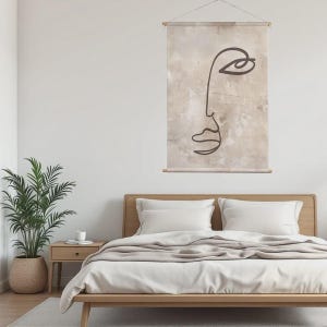 May include: A minimalist wall hanging featuring a line drawing of a face. The artwork is in dark brown against a beige background with a textured appearance. The piece is suspended by a wooden rod and string, above a wooden bed frame.