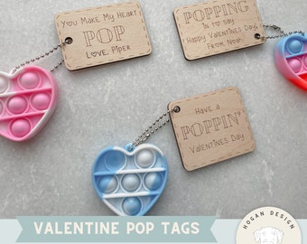 Puede incluir: Etiquetas de pop-it para el Día de San Valentín con un juguete pop-it en forma de corazón. Las etiquetas son de madera y tienen un diseño cortado con láser. El texto en las etiquetas dice "You make my heart pop", "Poppin' in to say Happy Valentines Day! From Noah", y "Have a Poppin' Valentines Day".