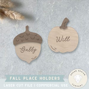 May include: Two wooden fall-themed place holders, one shaped like an acorn with "Gabby" engraved, and the other a pumpkin with "Will". The acorn top is a darker brown. The text "FALL PLACE HOLDERS" is at the bottom.