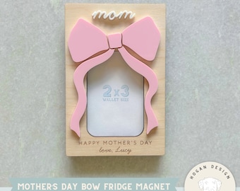 Op de afbeelding: Een houten fotolijst met een roze strik en het woord "mom" erop. De lijst is ontworpen om een ​​2x3 inch foto in portefeuilleformaat te bevatten. De tekst "Happy Mother's Day love, Lucy" is in de lijst gegraveerd.